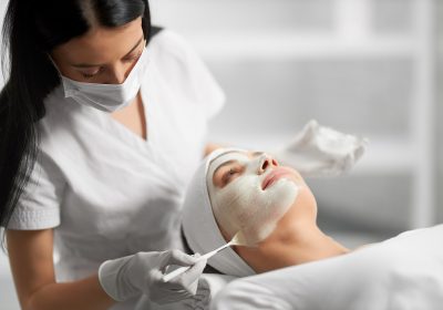 Side view of beautiful brunette beautician applying special white cream for face young cute patient. Concept of process procedure for improving face skin with cosmetics.